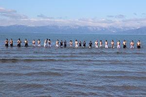 雪の積もる山を背に一列に並んで琵琶湖に入る学生たち